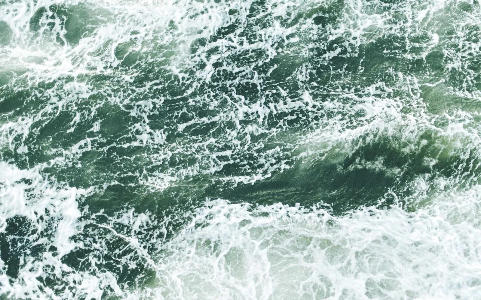 An aerial close-up of green ocean waves and white foam